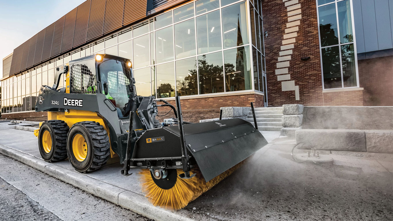 Chargeuse à direction différentielle John Deere équipée de l’accessoire balai orientable qui balaie le trottoir devant un commerce